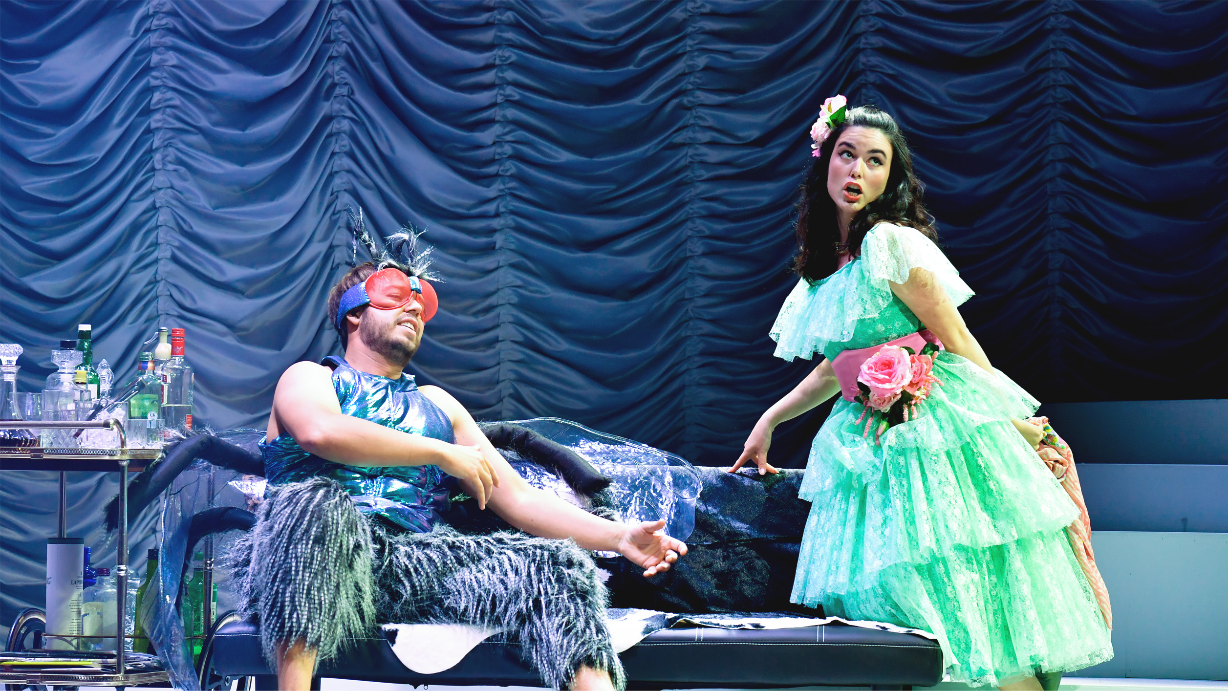 Two students performing in an opera, the man dressed as a fly sitting on a sofa, with a female student dressed in a light green, frilly dress, standing next to him, singing towards the audience, with a dark background.